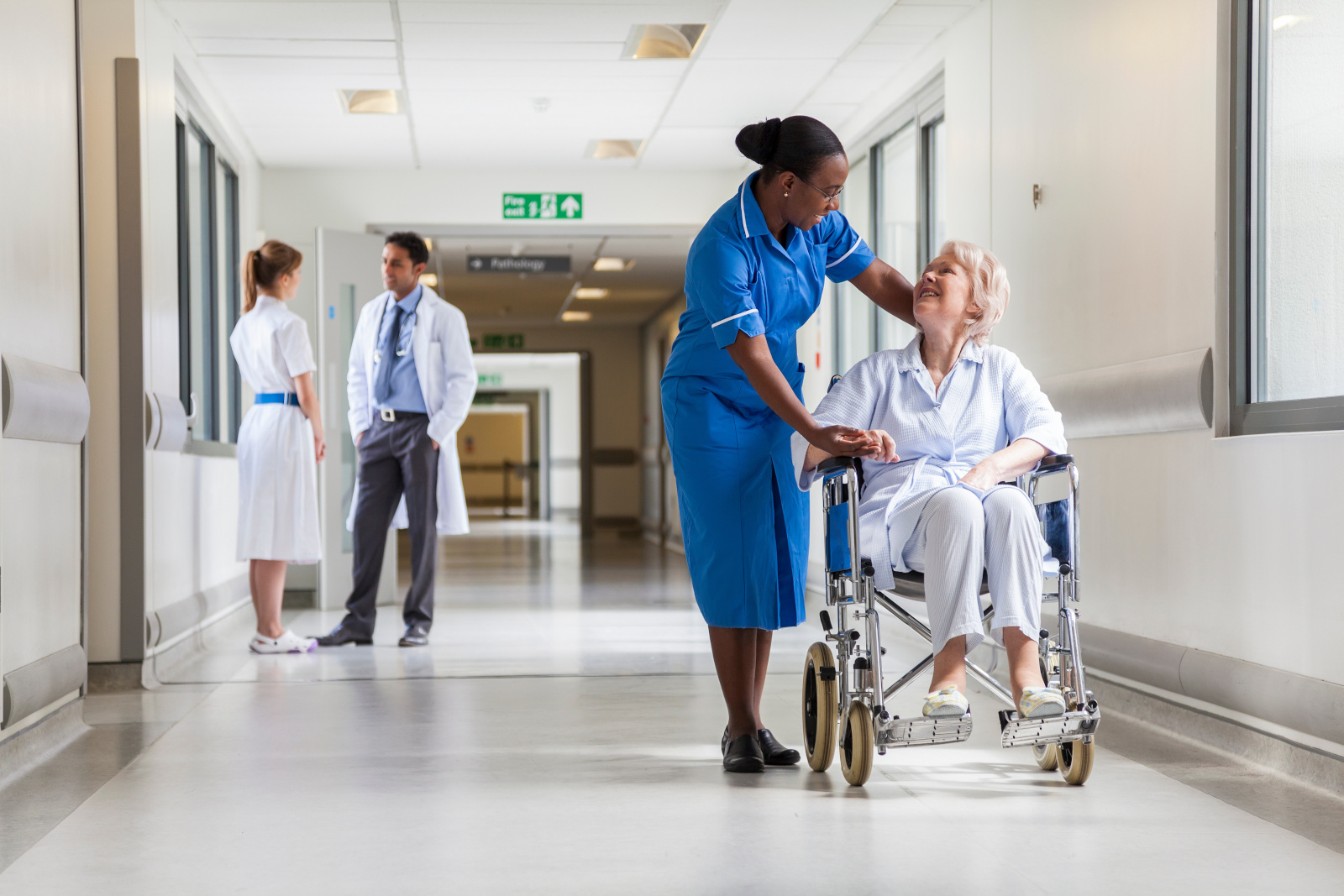 Senior Female Patient in Wheelchair & Nurse in Hospital Hospital corridor showing different roles working within a hospital. Temporary staff availability management with YouRecruit