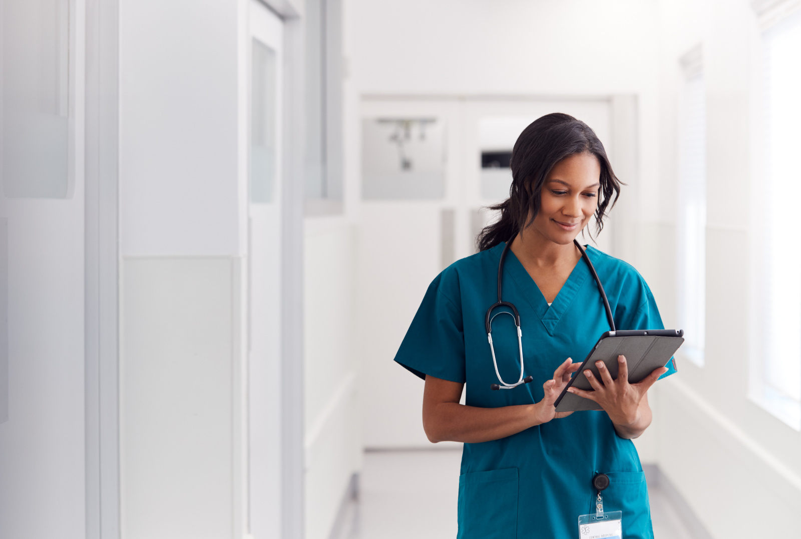 Female Doctor Wearing Scrubs In Hospital Corridor Using Digital Tablet Temporary nurse in hospital corridor checking her schedule and availability on the WorkTracker workshift appointments app