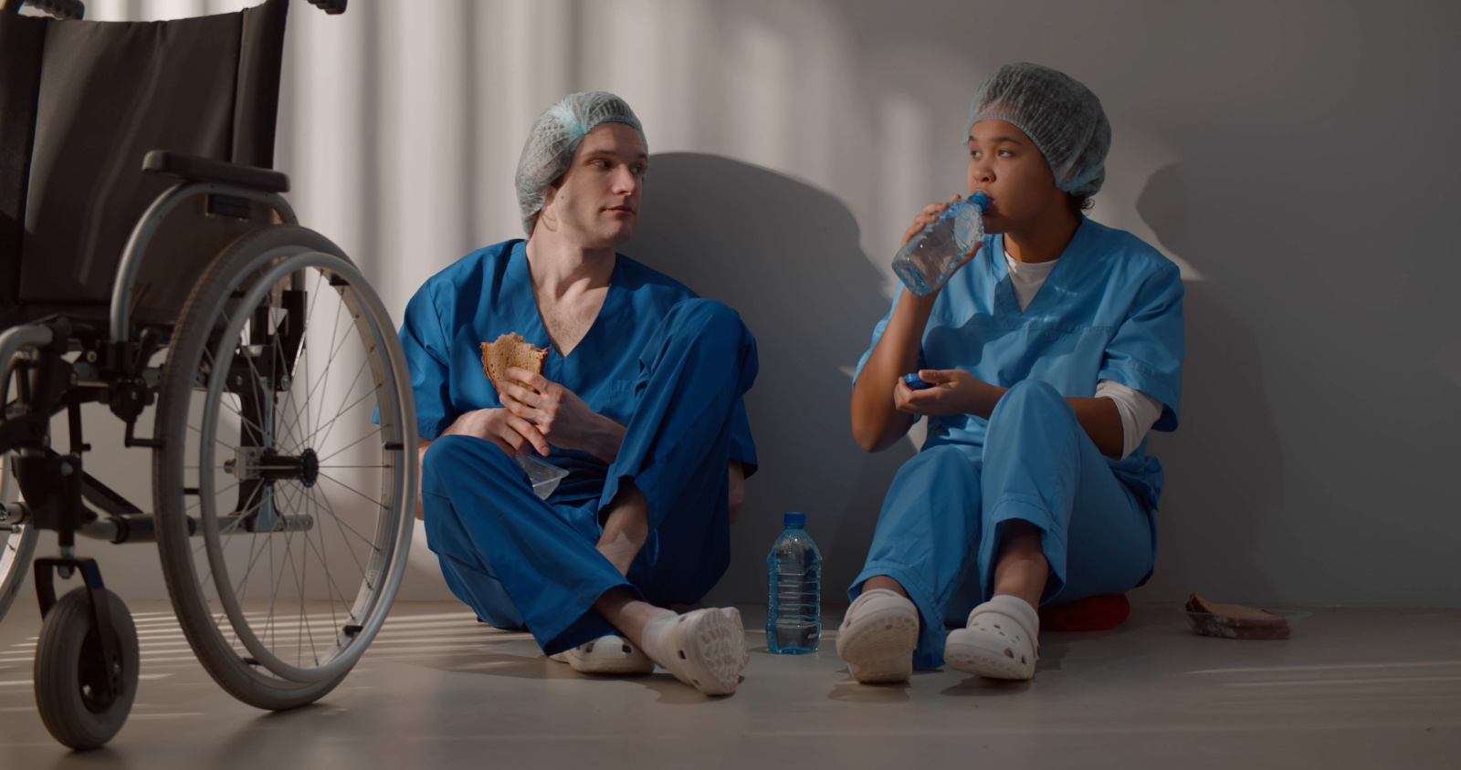 Diverse doctors eating and relaxing sitting on floor in hospital corridor Scheduled breaks in healthcare with WorkTracker mobile timesheets