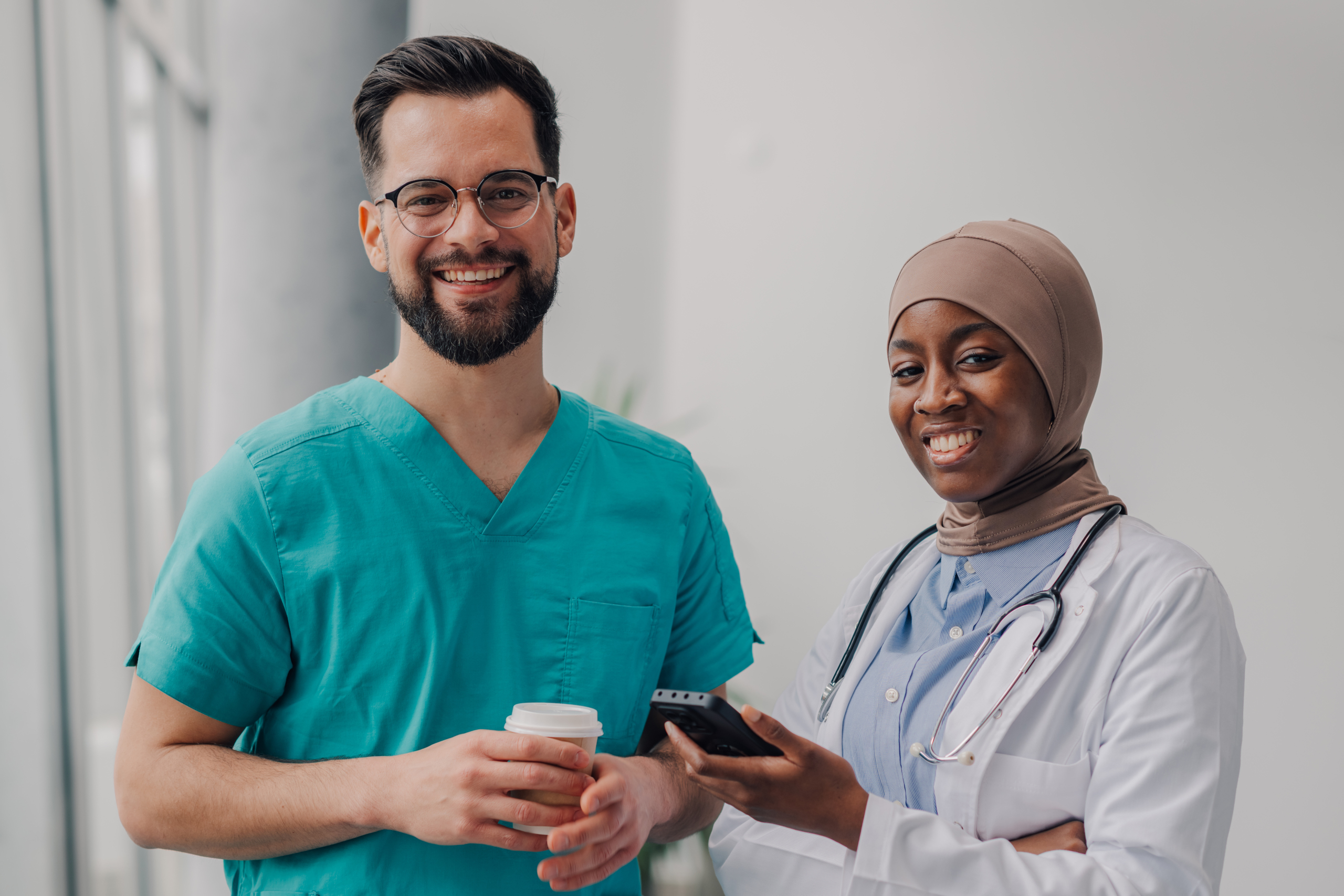 env-yr-doctors-smiling-and-holding-coffee-and-mobile-phon-2025-06-05-20-49-12-utc2 Male and female temporary doctors in healthcare setting on a break, female holding smartphone: The WorkTracker staff rota app