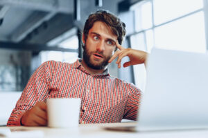 Puzzled looking recruitment consultant sat at desk with laptop and coffee cup: The YouRecruit healthcare software solution.