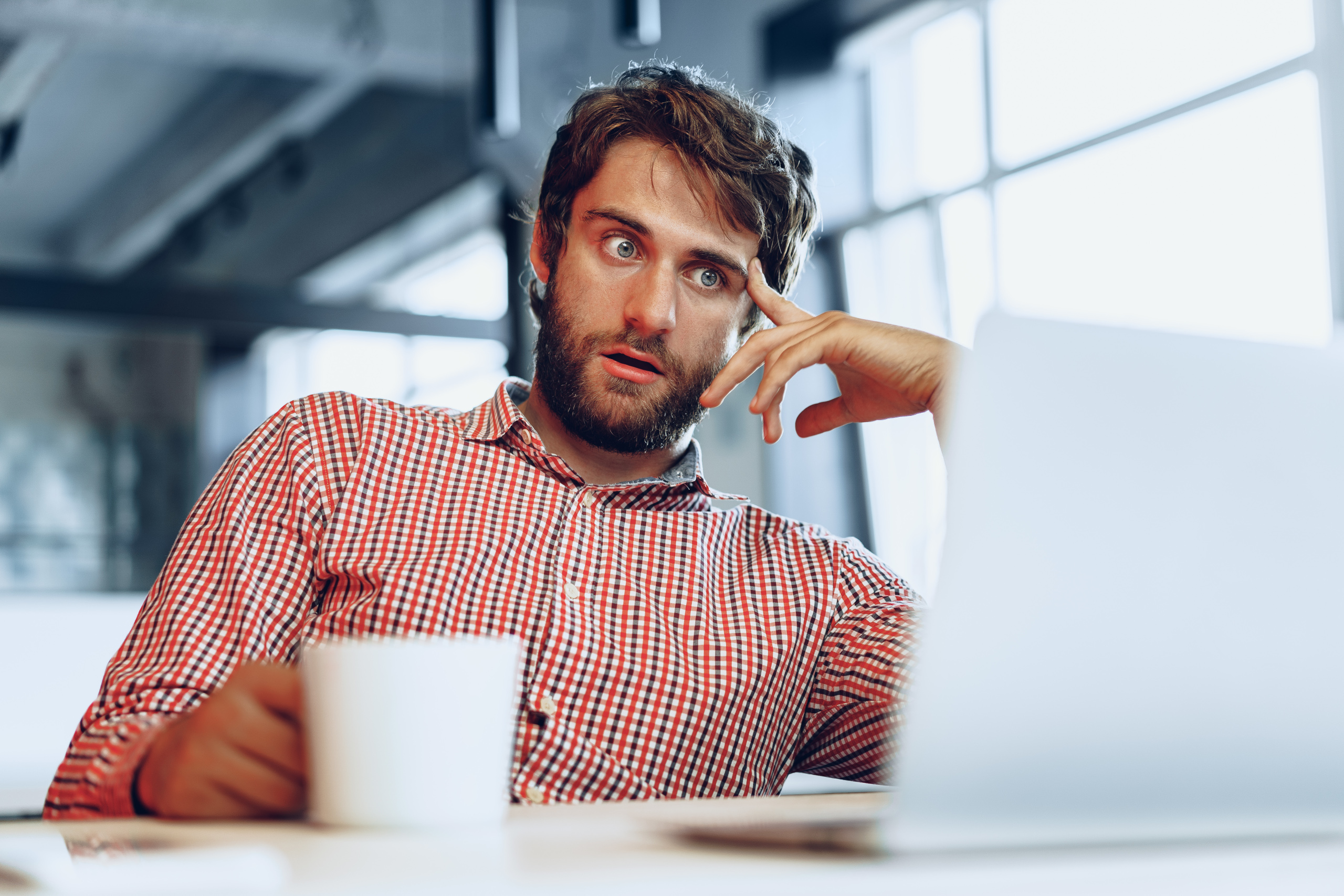 Puzzled looking recruitment consultant sat at desk with laptop and coffee cup: The YouRecruit healthcare software solution.